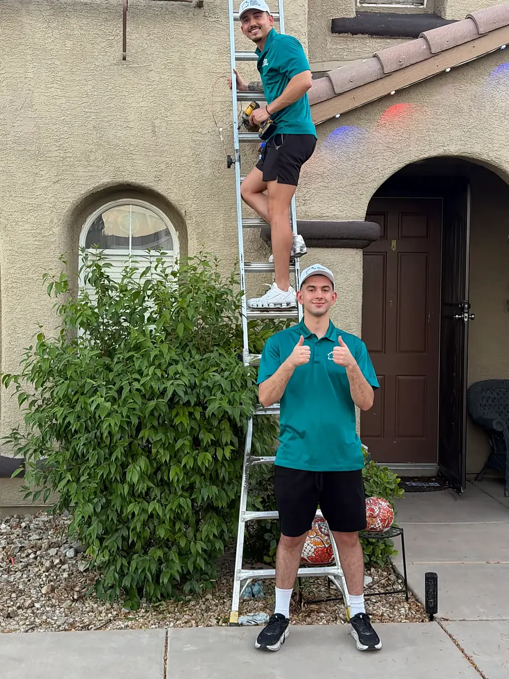 A Magic Season Lighting installer on a ladder with a drill while a teammate gives two thumbs up