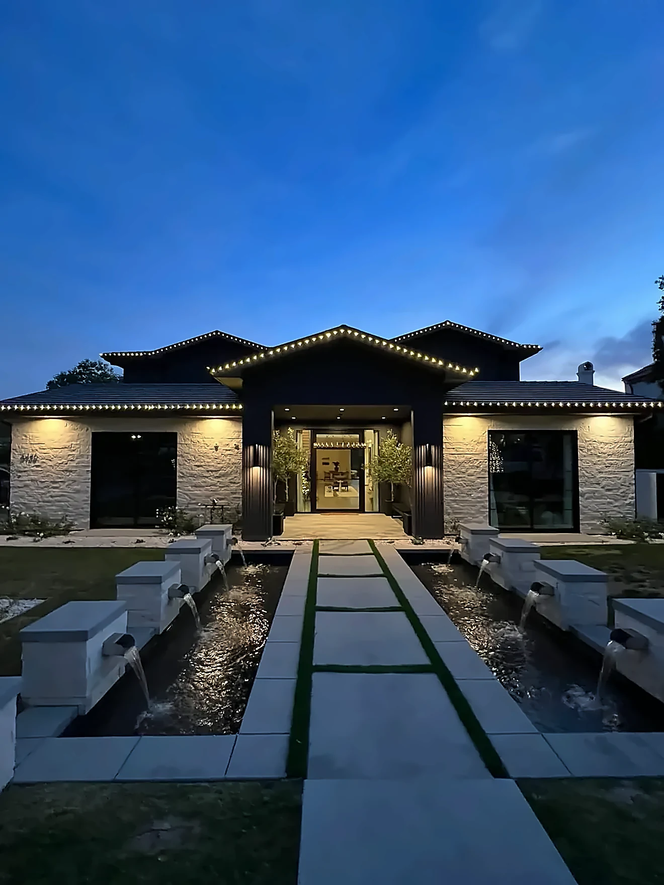 Modern home at dusk traced in warm white roofline lights above a lit fountain walkway